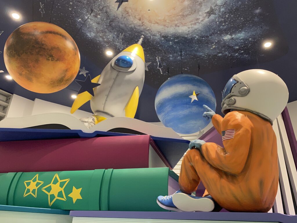 Figure of an astronaut pointing to a space scene of planets and a rocket. Oversized books are piled in front of the astronaut