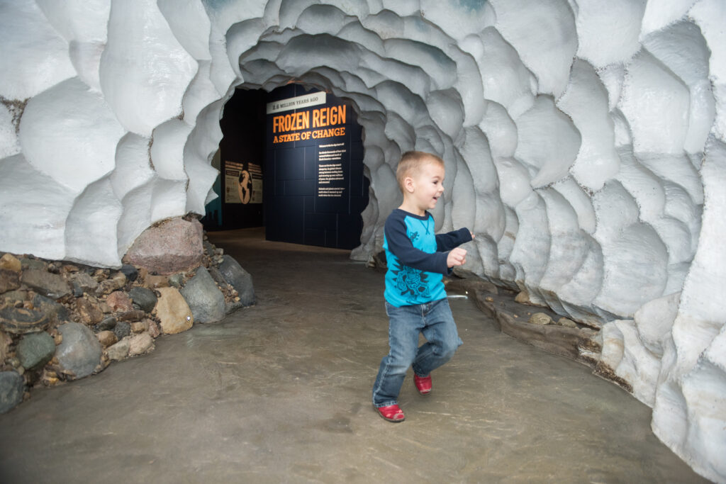 Happy child in front of white sculpted walls resembling ice. In the background a sign reading "frozen reign, a state of change" is shown.
