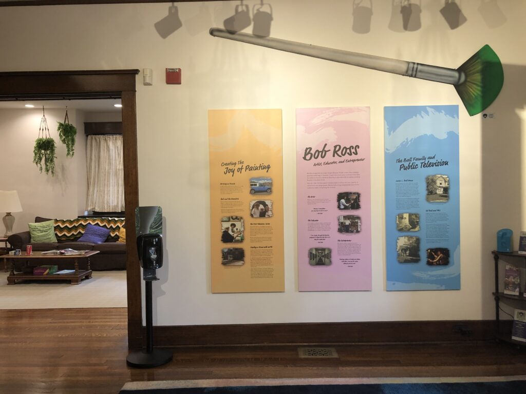 an introduction to the main exhibit shares information about Bob Ross, the Joy of Painting Show, and Public television with a giant paint brush graphic on the wall. Peaking to the left is a themed living room couch and coffee table.