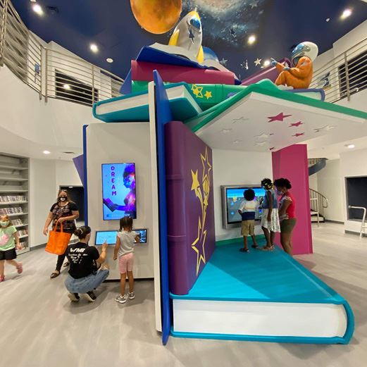 People engaging with interactive screens inside oversized books open at different angles to create a structure. On top of the structure is an astronaut and space theme.