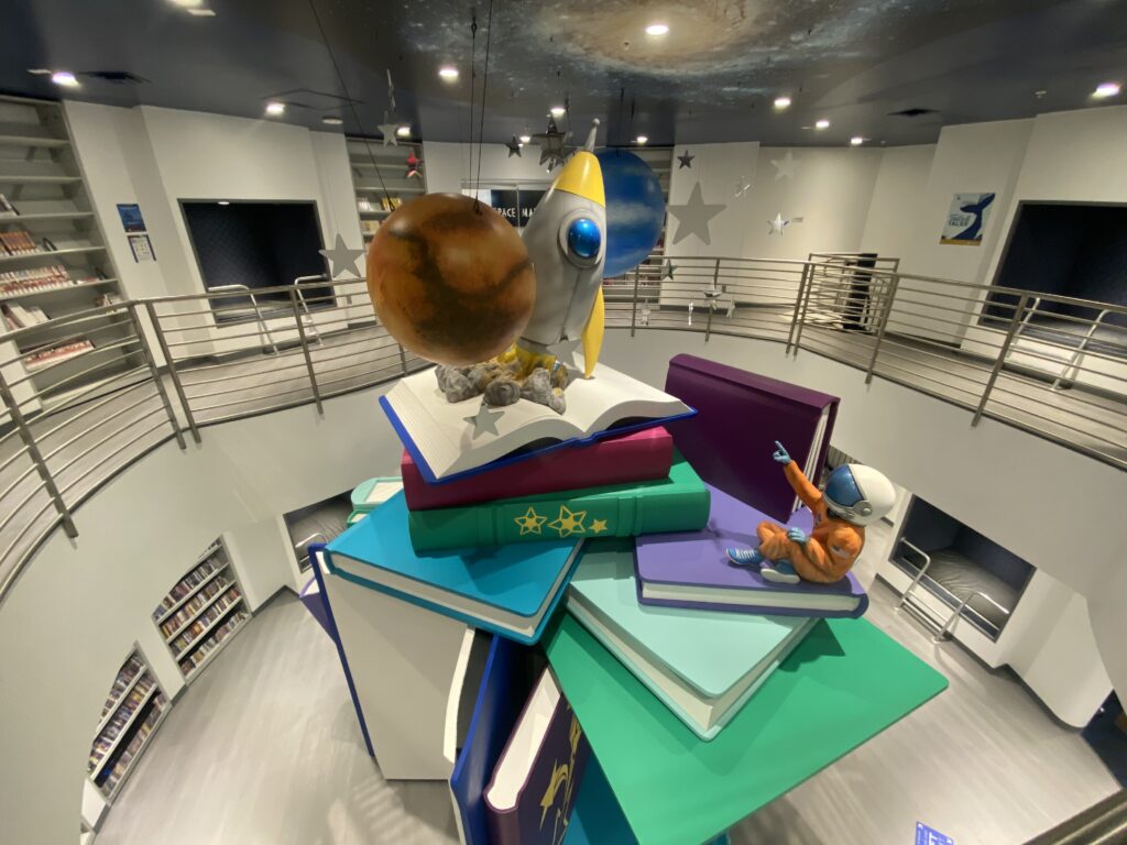 Aerial view of an astronaut pointing to a space scene featuring planets and a rocket. Oversized books are piled in front of the astronaut