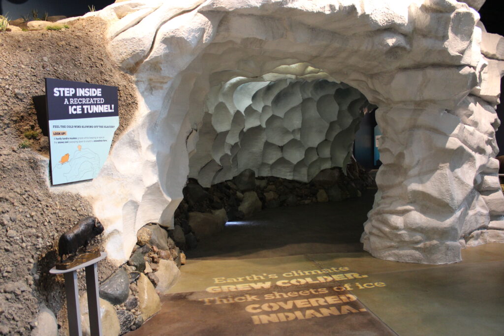 Entrance view of an ice cave with a sign to the left stating "step inside a recreated ice tunnel" and words on the floor that say "Earth's climate grew colder. Thick sheets of ice covered Indiana."