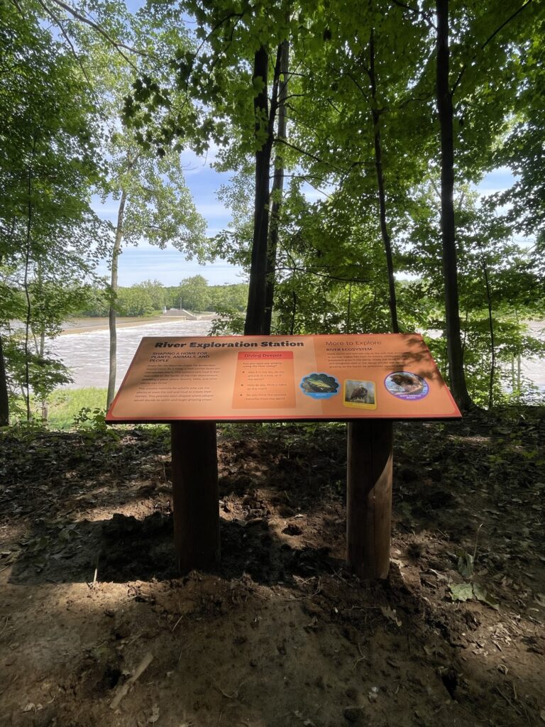 An exterior exhibit sign explaining a river exploration station set in the woods looking out to the water