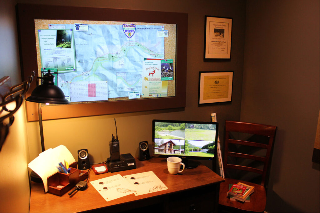 replica of a ranger's station including a computer screen showing multiple areas of the park, a billboard with paperwork, and an assortment of office clutter on the desk