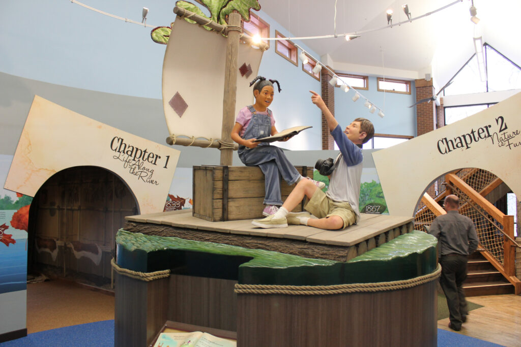 Models of two children sitting on a raft with a sail. One child is reading a book and the other is holding a camera and pointing. In the background you see an entry way with "Chapter 1 Life along the river" over the doorway. There is another doorway with the title "Chapter 2 Nature is Fun."